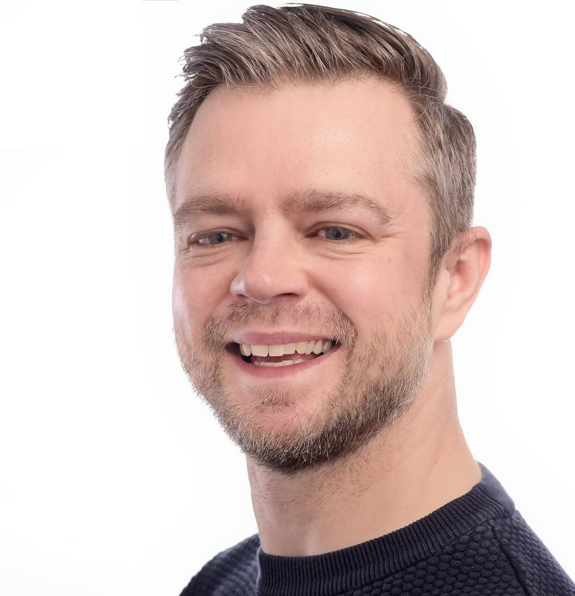 A man smiling towards the camera wearing a blue jumper, against a white background.