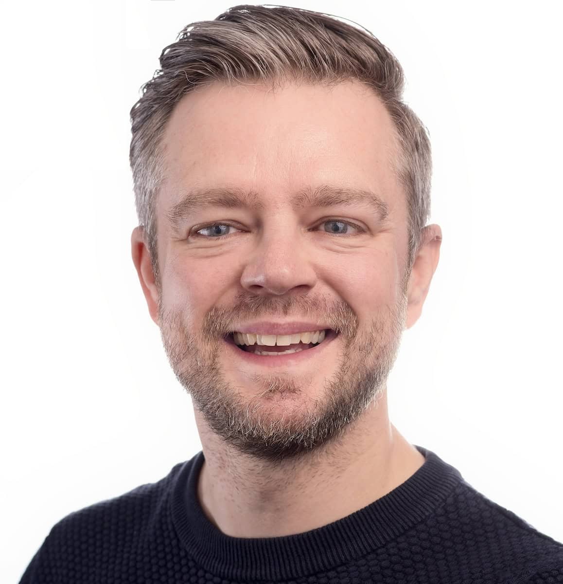 A man smiling straight on towards the camera wearing a blue jumper, against a white background.